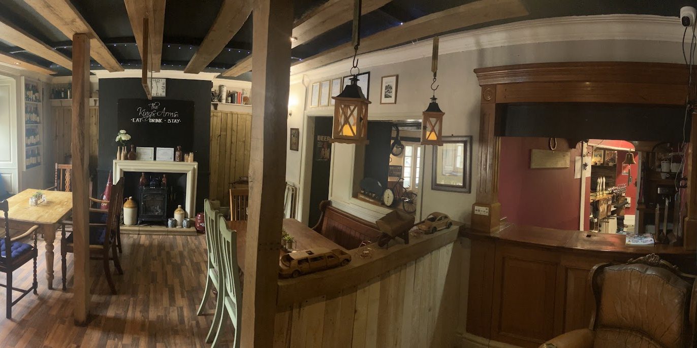 Inside The Kings Arms: exposed beams, a log burner, a chalkboard reading Eat Drink Stay, and the wooden bar beyond.